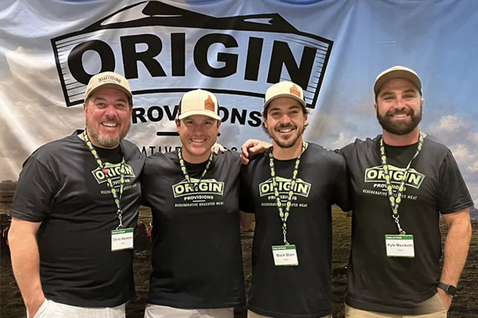 Four Origin Provisions founders standing together in front of the Origin Provisions banner